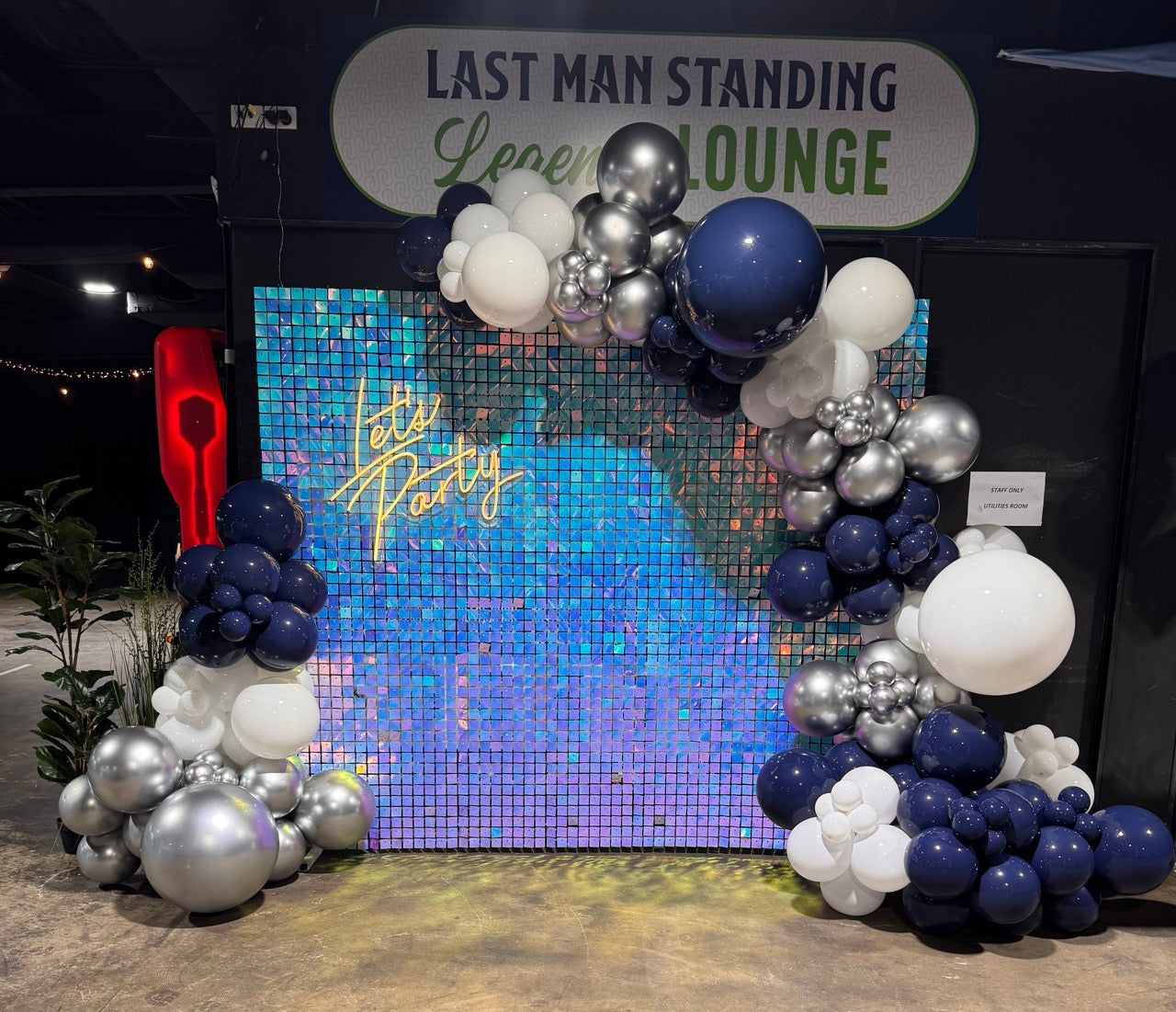 Decorative balloon arch with 'Let's Party' text in front of a colorful mosaic wall with 'Last Man Standing Lounge' sign.