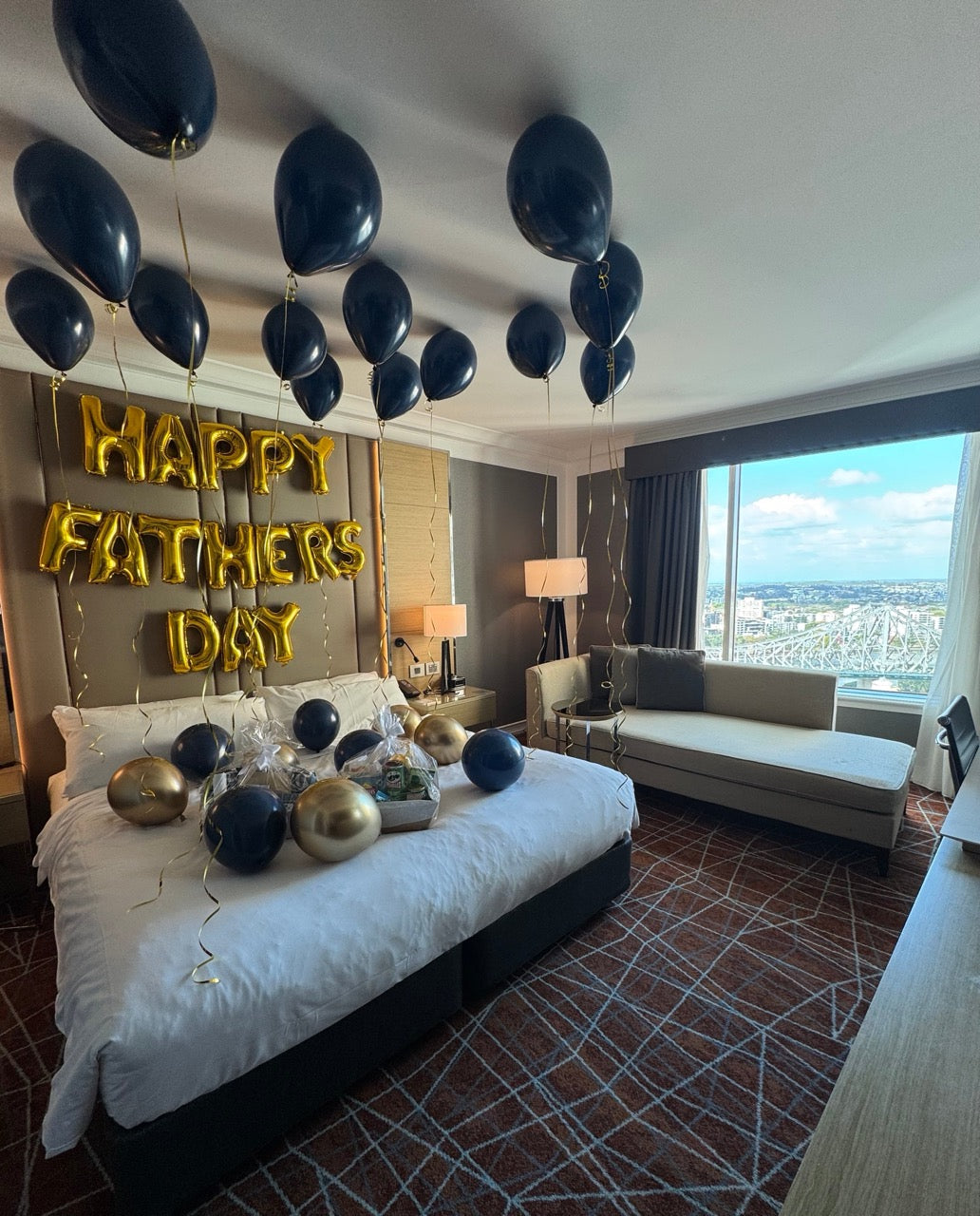Decorated bedroom with balloons and 'Happy Father's Day' sign, overlooking a cityscape.