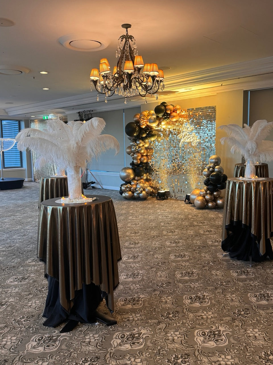Decorated event space with palm tree sculptures, balloons, and a chandelier.
