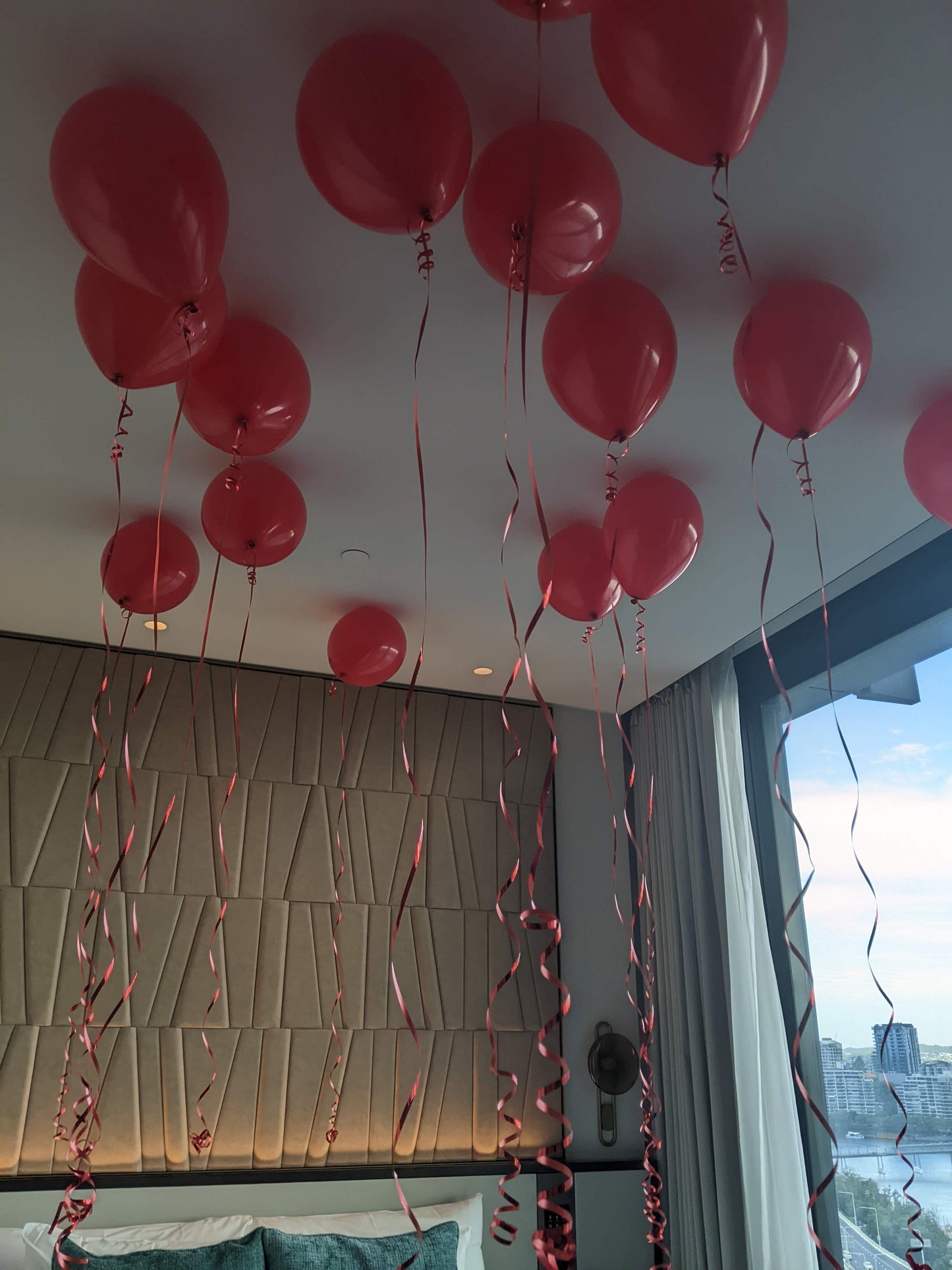 Valentine's Day balloons with ribbons on a ceiling with a cityscape view outside a window.