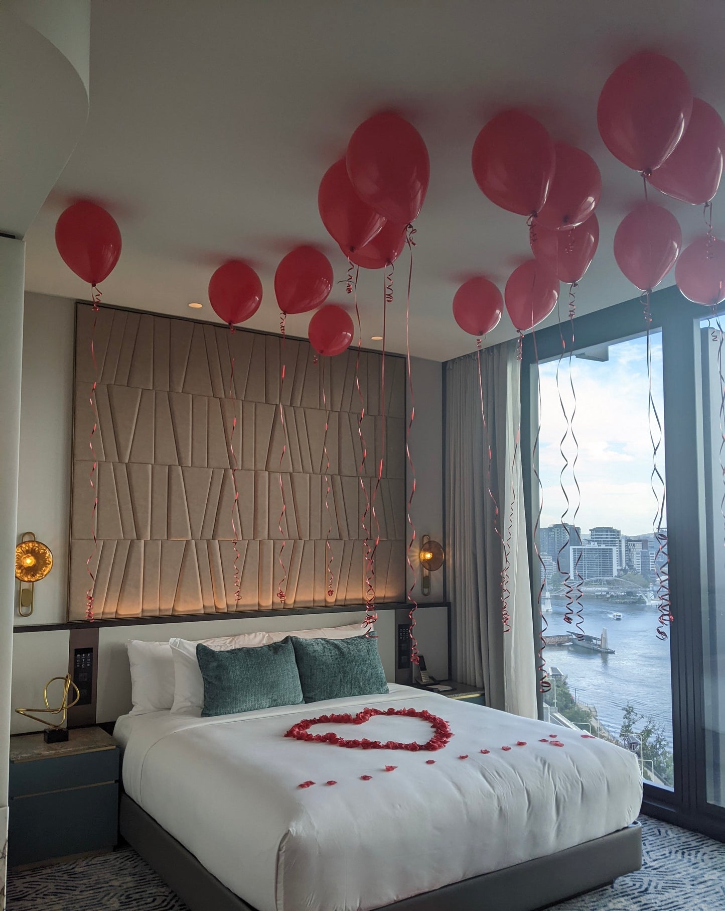 Decorated bedroom with balloons and heart-shaped rose petals on a bed, overlooking a cityscape.