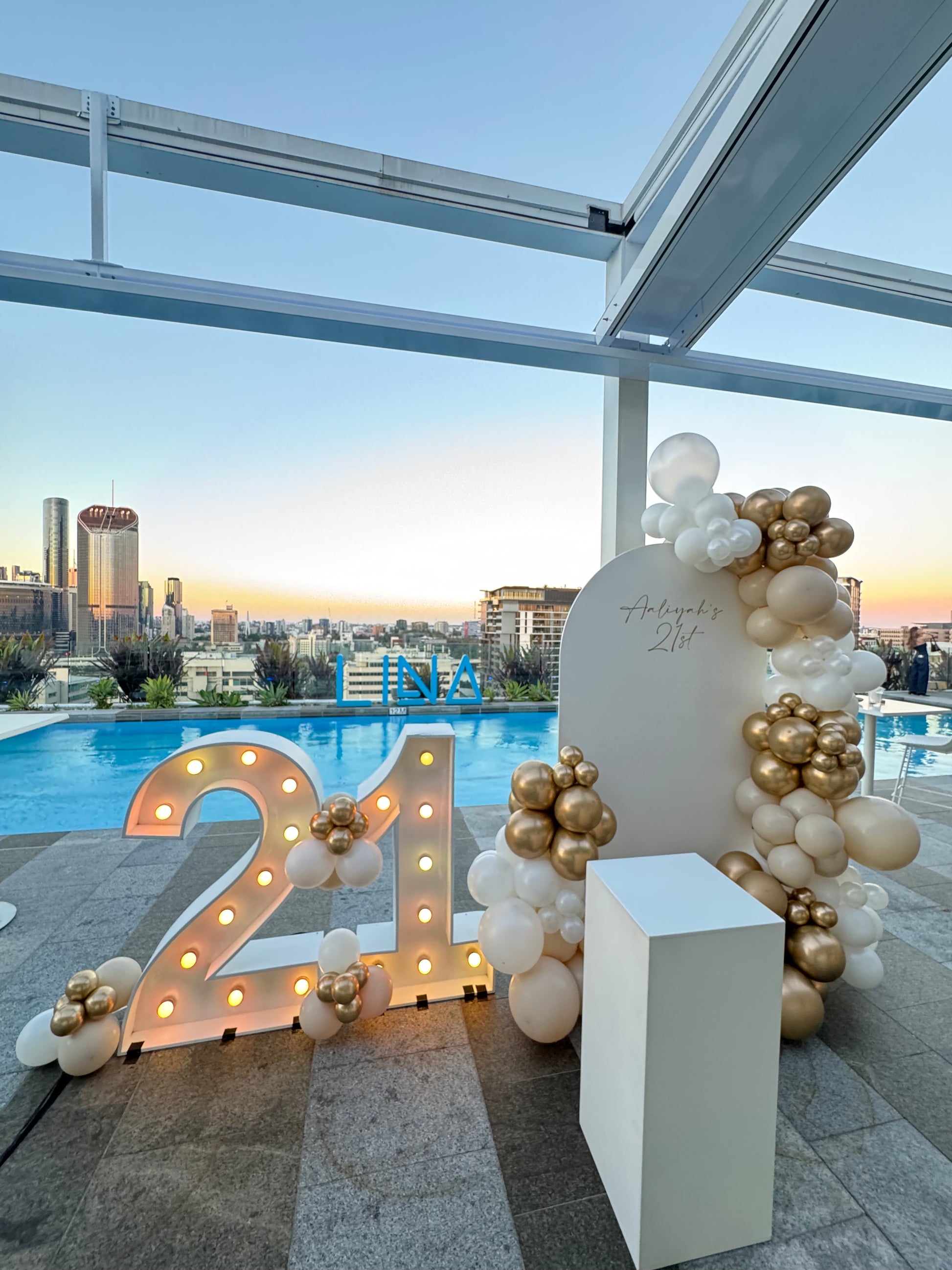 Light up number balloon arch with florals, cake plinth and personalised signage at a Brisbane birthday event, rooftop bar