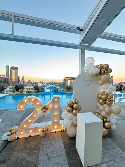 Light up number balloon arch with florals, cake plinth and personalised signage at a Brisbane birthday event, rooftop bar