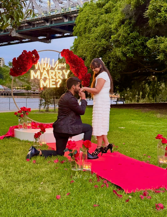 Man proposing to a woman with 'Will You Marry Me?' sign in a park setting.